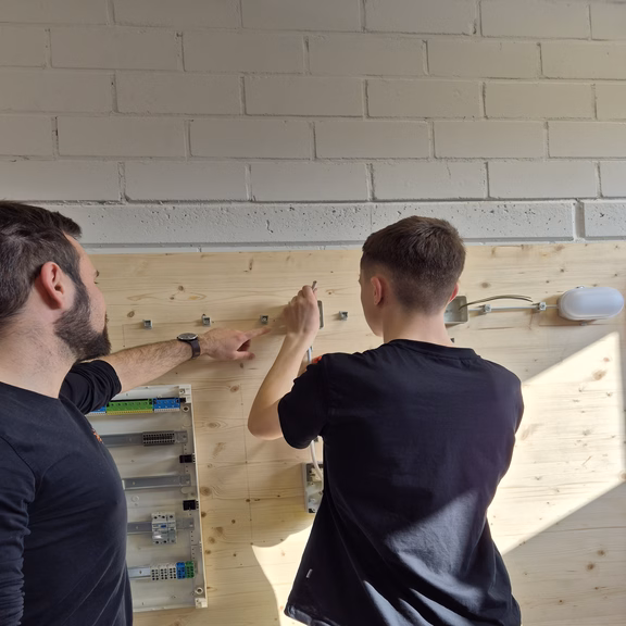 Ausbilder erklärt einem Auszubildenden den Aufbau einer elektrischen Schaltung an einer Übungswand in der Azubiwerkstatt.