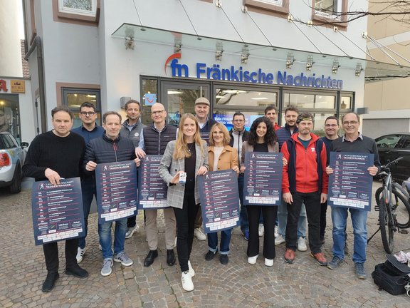 Laufgemeinschaft Tauberfranken Gruppe von Menschen steht vor dem Gebäude der „Fränkische Nachrichten“ und hält Plakate zur Volkslaufserie der Laufgemeinschaft Tauberfranken in die Kamera; Außenaufnahme auf einem gepflasterten Platz.