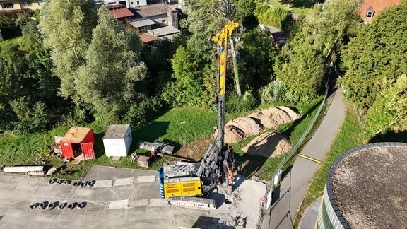 Bauarbeiten mit einem über 40 Tonnen schweren Bohrgerät auf einer Baustelle neben einem Gehweg und mehreren Containern, umgeben von Bäumen und Wohnhäusern.