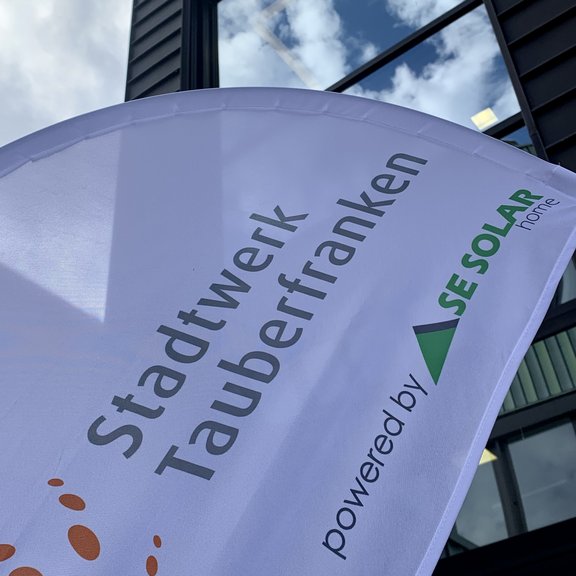 Ein weißes Werbesegel mit der Aufschrift „Stadtwerk Tauberfranken powered by SE SOLAR home“ weht im Wind vor einem modernen Gebäude mit großen Glasfenstern, in denen sich der blaue Himmel mit Wolken spiegelt.