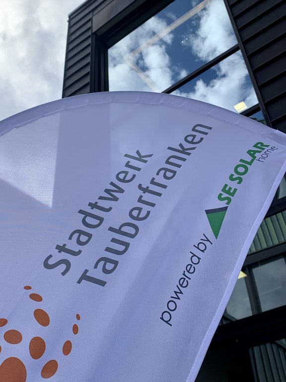 Ein weißes Werbesegel mit der Aufschrift „Stadtwerk Tauberfranken powered by SE SOLAR home“ weht im Wind vor einem modernen Gebäude mit großen Glasfenstern, in denen sich der blaue Himmel mit Wolken spiegelt.