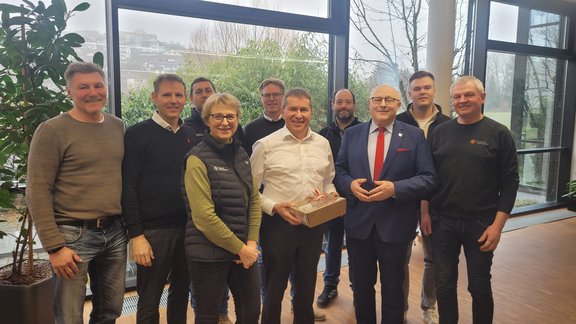 Eine Gruppe von neun Männern und einer Frau steht in einem hellen Raum mit großen Fenstern, durch die grüne Sträucher zu sehen sind. Die Personen lächeln freundlich in die Kamera. Eine Person in der Mitte hält ein Geschenk mit roter Schleife, während ein Mann im Anzug und roter Krawatte daneben steht. In dem Raum ist auf der linken Seite eine grüne Pflanze zu sehen..
