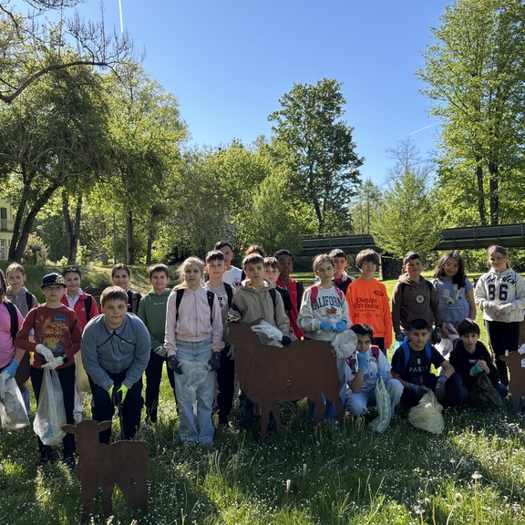 Eine Gruppe von Kindern steht auf einer Wiese im Grünen. Die Kinder tragen teilweise Arbeitshandschuhe und halten Müllsäcke. Im Hintergrund sind Bäume, eine Brücke und ein sonniger Parkbereich zu sehen.