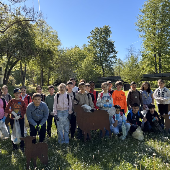 Müllsammelaktion Grundschule 2026 Eine Gruppe von Kindern steht auf einer Wiese im Grünen. Die Kinder tragen teilweise Arbeitshandschuhe und halten Müllsäcke. Im Hintergrund sind Bäume, eine Brücke und ein sonniger Parkbereich zu sehen.