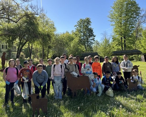 Eine Gruppe von Kindern steht auf einer Wiese im Grünen. Die Kinder tragen teilweise Arbeitshandschuhe und halten Müllsäcke. Im Hintergrund sind Bäume, eine Brücke und ein sonniger Parkbereich zu sehen.