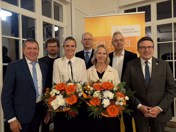 Es zeigt eine Gruppe von sieben Personen bei einer Veranstaltung, zwei davon mit großen Blumensträußen. Im Hintergrund ist ein Banner mit dem gut erkennbaren Schriftzug „Stadtwerk Tauberfranken“ zu sehen.
