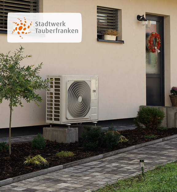 Außeneinheit einer Wärmepumpe vor einem modernen Wohnhaus mit Vorgarten und gepflastertem Weg. In der oberen linken Ecke befindet sich das Logo des Stadtwerks Tauberfranken.