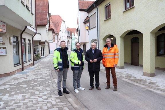 Vier Personen stehen auf einer frisch gepflasterten Straße in einer Altstadt. Die Männer tragen wetterfeste Kleidung, zwei in neongelben Jacken und einer in einer orangefarbenen Warnjacke. Im Hintergrund sind enge Gassen und historische Gebäude mit pastellfarbenen Fassaden zu sehen. Die Szene vermittelt den Eindruck einer Ortsbesichtigung oder Abnahme von Bauarbeiten.
