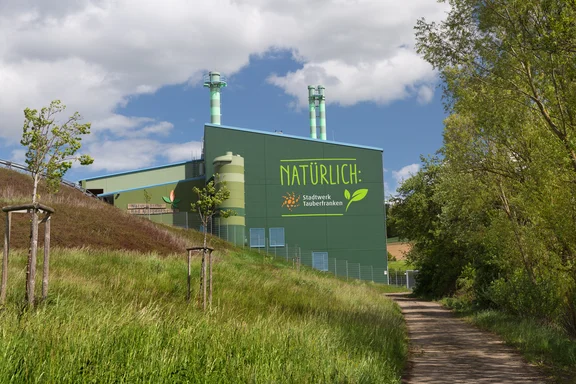 Ein modernes, grünes Gebäude mit der Aufschrift „Naturwärme Bad Mergentheim“ steht unter blauem Himmel. Im Vordergrund ist eine Zufahrtsstraße und links und rechts sind satt grün Bäume zu sehen. Das Bild vermittelt den Eindruck von Nachhaltigkeit und umweltfreundlicher Energieerzeugung.