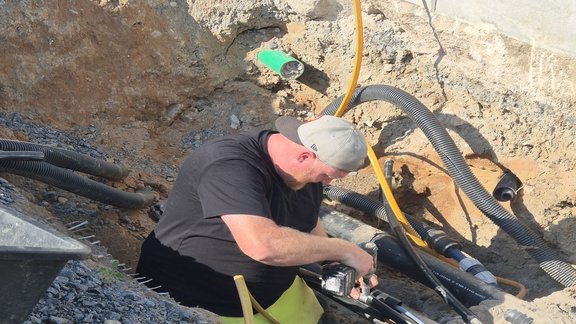 Ein Arbeiter sitzt in einer Baugrube und führt Montagearbeiten an einem dicken schwarzen Kabel durch. Er trägt ein schwarzes T-Shirt, neon-gelbe Arbeitshose und eine graue Basecap. Um ihn herum liegen verschiedene Werkzeuge, Rohre und Kabel in der Erde. Die Szene spielt sich an einem sonnigen Tag ab.