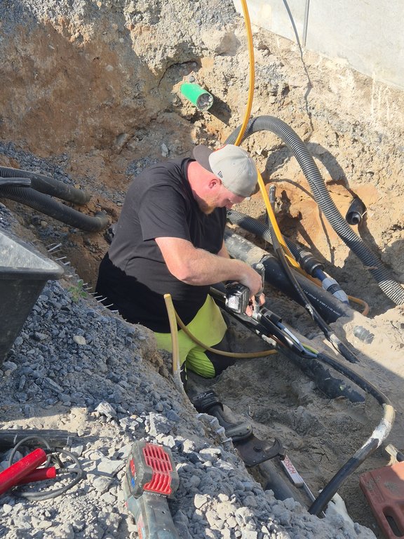 Ein Arbeiter sitzt in einer Baugrube und führt Montagearbeiten an einem dicken schwarzen Kabel durch. Er trägt ein schwarzes T-Shirt, neon-gelbe Arbeitshose und eine graue Basecap. Um ihn herum liegen verschiedene Werkzeuge, Rohre und Kabel in der Erde. Die Szene spielt sich an einem sonnigen Tag ab.