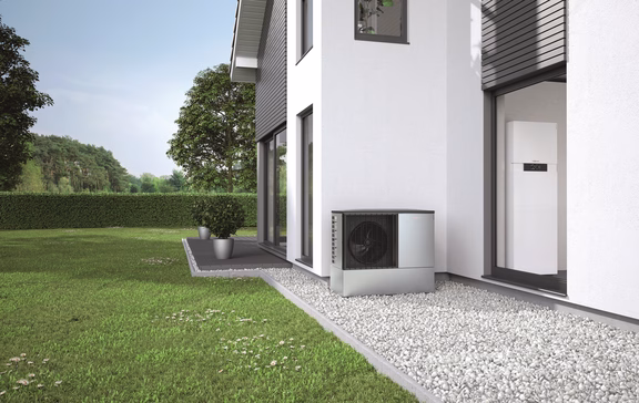 Wärmepumpe Moderne Luft-Wasser-Wärmepumpe der Marke Viessmann im Garten eines Einfamilienhauses, installiert auf einem Kiesbett direkt neben der Hauswand. Im Hintergrund sichtbar: großzügige Rasenfläche, Hecke und Bäume.