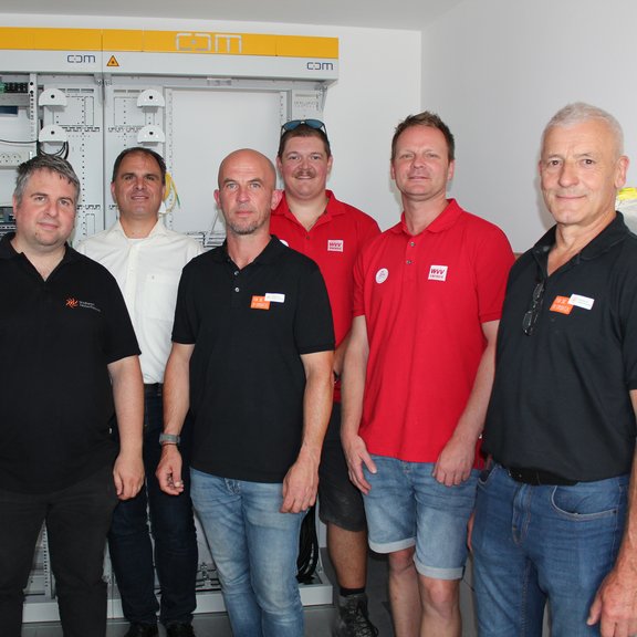 Auf dem Bild sind 7 Männer vor einem Schaltschrank zu sehen. Einige tragen Hemden ein paar auch T-Shirts mit Firmenlogo darauf.