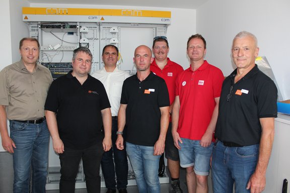 Auf dem Bild sind 7 Männer vor einem Schaltschrank zu sehen. Einige tragen Hemden ein paar auch T-Shirts mit Firmenlogo darauf.