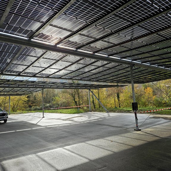 Parkplatz-PV-Anlage Ein Auto steht auf einer Parkplatzfläche, die mit PV-Modulen überdacht ist. Die Sonne scheint durch die Paneele und es ist ein freundlicher Wintertag.