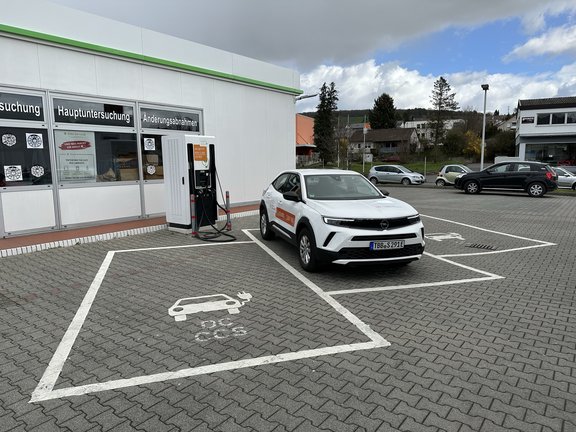 Ein weißer Opel Mokka-e steht an einer E-Ladesäule auf einem Parkplatz. Die Schnellladestation befindet sich vor einem Gebäude mit Schildern für Hauptuntersuchung und Änderungsabnahmen. Der Himmel ist teils bewölkt, und im Hintergrund sind mehrere geparkte Autos sowie Häuser mit Gärten zu sehen.