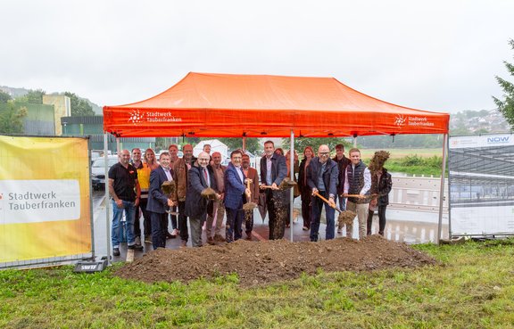 Das Bild vermittelt den Eindruck eines Spatenstichs für ein Projekt. Unter einem orangenen Pavillion mit dem Logo des Stadtwerks Tauberfranken stehen viele Menschen. Im Vordergrund stehen 6 Männer mit einem Spaten vor einem Erdhügel und werfen Erde in die Luft. Die Männer tragen Anzug und es regnet.