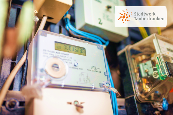 Nahaufnahme eines Smart Meters mit sichtbarem Zählerstand, umgeben von elektrischen Leitungen und weiteren Bauteilen. Rechts oben ist das Logo von Stadtwerk Tauberfranken eingeblendet.