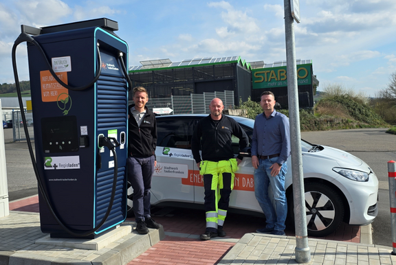 Drei Männer stehen neben einer öffentlichen Schnellladestation für Elektroautos und einem weißen E-Auto mit dem Logo des Stadtwerks Tauberfranken. Die Ladestation trägt den Schriftzug „Aufladen mit Heimatstrom von hier“. Im Hintergrund ist ein Gewerbegebiet mit einem Baumarkt zu sehen. Der Himmel ist teils bewölkt.