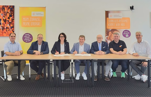Kooperation Stadtwerk und Telekom Sieben Personen sitzen an einem Tisch bei der Unterzeichnung einer Kooperationsvereinbarung. Im Hintergrund stehen zwei Roll-up-Banner des Stadtwerks Tauberfranken zu Energie- und Internetangeboten.