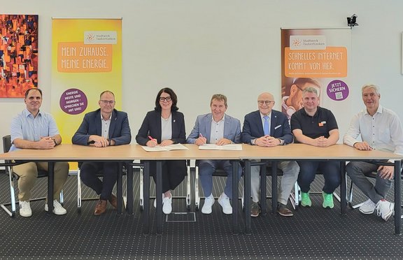 Kooperation Stadtwerk und Telekom Sieben Personen sitzen an einem Tisch bei der Unterzeichnung einer Kooperationsvereinbarung. Im Hintergrund stehen zwei Roll-up-Banner des Stadtwerks Tauberfranken zu Energie- und Internetangeboten.