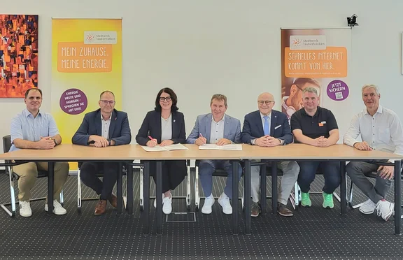 Sieben Personen sitzen an einem Tisch bei der Unterzeichnung einer Kooperationsvereinbarung. Im Hintergrund stehen zwei Roll-up-Banner des Stadtwerks Tauberfranken zu Energie- und Internetangeboten.