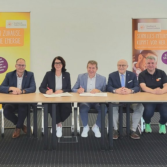 Kooperation Stadtwerk und Telekom Sieben Personen sitzen an einem Tisch bei der Unterzeichnung einer Kooperationsvereinbarung. Im Hintergrund stehen zwei Roll-up-Banner des Stadtwerks Tauberfranken zu Energie- und Internetangeboten.