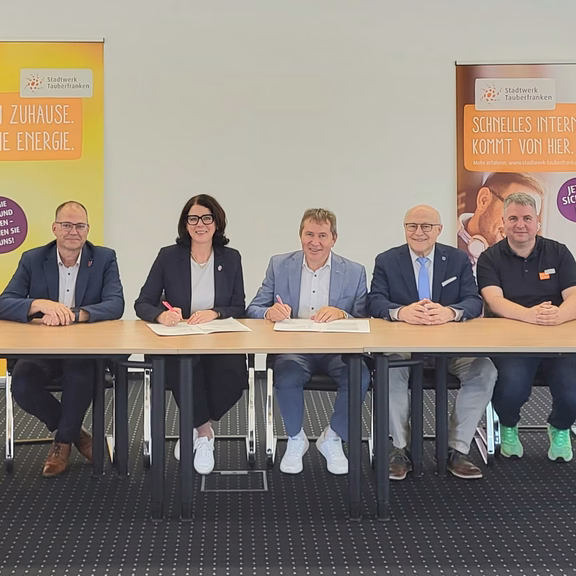Kooperation Stadtwerk und Telekom Sieben Personen sitzen an einem Tisch bei der Unterzeichnung einer Kooperationsvereinbarung. Im Hintergrund stehen zwei Roll-up-Banner des Stadtwerks Tauberfranken zu Energie- und Internetangeboten.