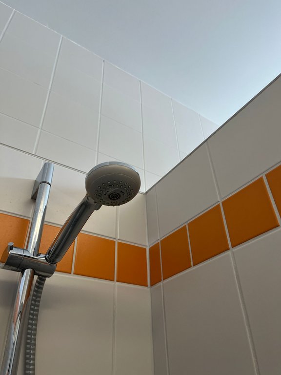 Nahaufnahme einer Dusche mit verchromtem Duschkopf, der an einer verstellbaren Halterung befestigt ist. Die Wände sind mit weißen Fliesen und einer dekorativen orangefarbenen Fliesenreihe versehen.