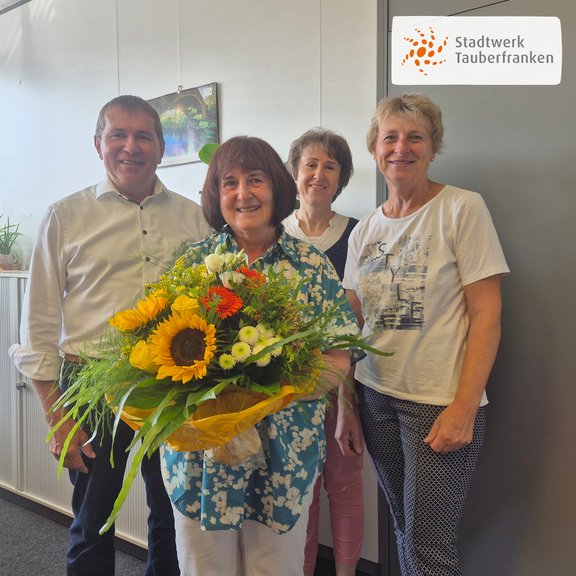 Ein Mann und 3 Frauen stehen in einem hellen, freundlichen Büroraum. Die Personen sind leger gekleidet. Eine Frau hält einen Blumenstrauß mit Sonnenblumen in der Hand. Die Szene vermittelt eine Gratulation zu einem Betriebsjubiläum.