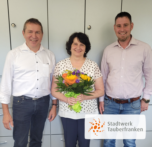Drei Personen stehen vor einem grauen Aktenschrank und lächeln in die Kamera. In der Mitte hält eine Frau mit dunkelbraunen Haaren einen bunten Blumenstrauß mit Sonnenblumen, Rosen und anderen Blüten. Links und rechts von ihr stehen zwei Männer in Hemd und Jeans. Unten rechts im Bild ist das Logo von „Stadtwerk Tauberfranken“ eingeblendet.