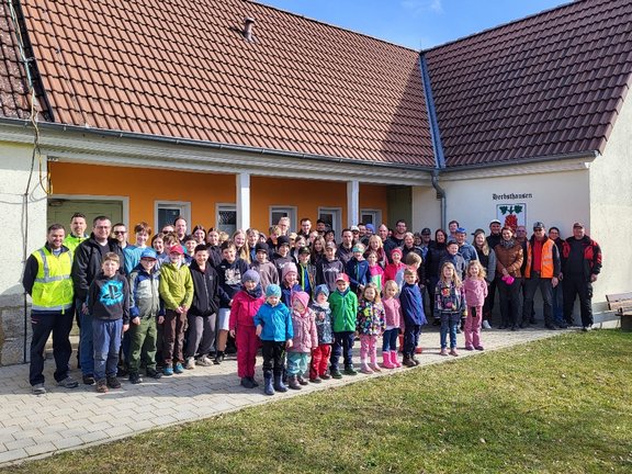 Viele Personen von jung bis alt stehen vor einem Gebäude und lächeln in die Kamera. Die Sonne scheint.