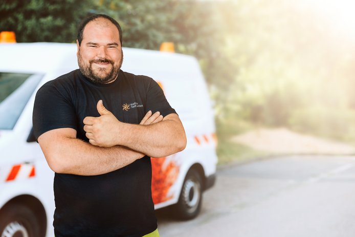 Ein Mann mittleren Alters im schwarzen T-Shirt mit Stadtwerk-Logo steht lächelnd mit verschränkten Armen vor einem weißen Baustellenbus. Im Hintergrund ist eine grüne Hecke zu sehen. 