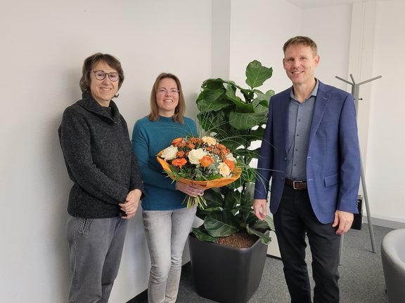 Drei Personen stehen nebeneinander in einem Büro. Die mittlere Person hält einen großen Blumenstrauß mit orange-weißen Blumen in der Hand. Im Hintergrund ist eine große grüne Zimmerpflanze zu sehen.