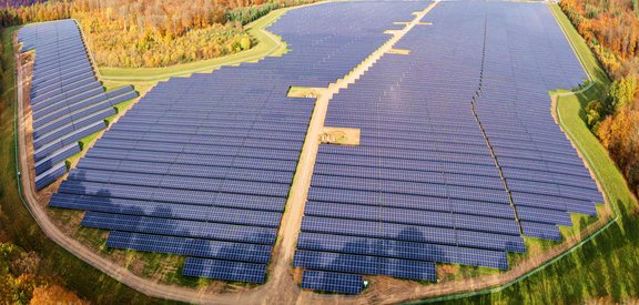 Luftaufnahme eines riesigen Solarparks mit unzähligen Solarpanelen, die sich über eine weite Fläche erstrecken. Ein sandiger Weg verläuft durch die Mitte der Anlage. Die Umgebung besteht aus grünen Wiesen und herbstlich gefärbten Wäldern mit gelben, orangen und roten Blättern.