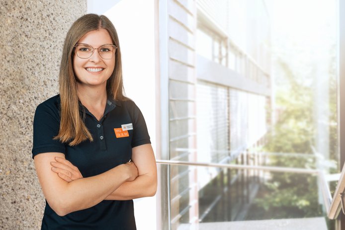 Eine freundlich lächelnde Frau mit langen braunen Haaren und Brille steht lächelnd mit verschränkten Armen im Flur eines Bürogebäudes. Sie trägt ein schwarzes T-Shirt mit Stadtwerk-Tauberfranken Logo.