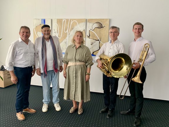 Fünf Personen stehen nebeneinander in einem Ausstellungsraum vor einem abstrakten Kunstwerk. Zwei Männer rechts halten jeweils ein Musikinstrument – eine Tuba und eine Posaune – und tragen weiße Hemden. In der Mitte steht eine Frau in einem beigen Kleid, links von ihr ein Mann mit Mütze und Schal, daneben ein weiterer Mann im weißen Hemd. Alle lächeln in die Kamera.