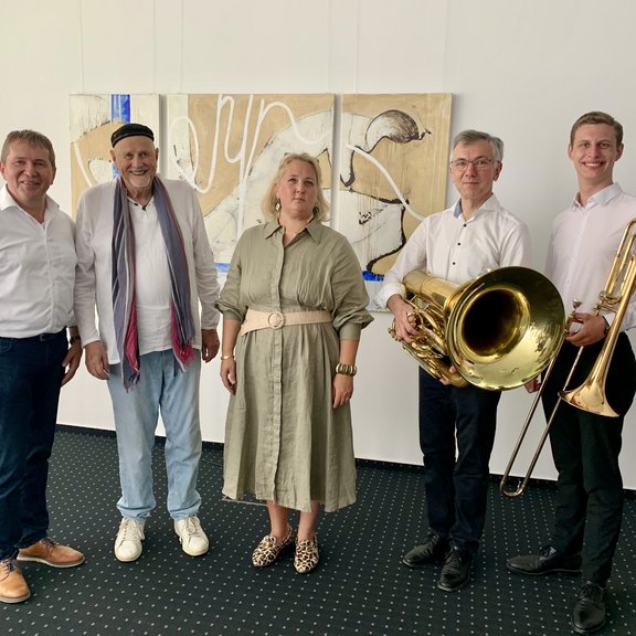 Fünf Personen stehen nebeneinander in einem Ausstellungsraum vor einem abstrakten Kunstwerk. Zwei Männer rechts halten jeweils ein Musikinstrument – eine Tuba und eine Posaune – und tragen weiße Hemden. In der Mitte steht eine Frau in einem beigen Kleid, links von ihr ein Mann mit Mütze und Schal, daneben ein weiterer Mann im weißen Hemd. Alle lächeln in die Kamera.