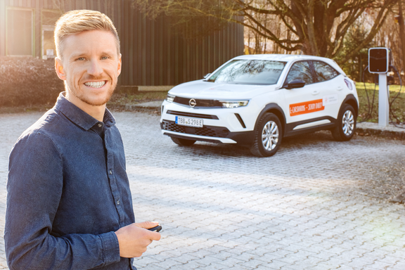Mann mit kurzem blondem Haar und blauem Hemd steht lächelnd vor einem weißen Opel SUV mit orangefarbenem Logo. Im Hintergrund ist eine Ladesäule sichtbar.