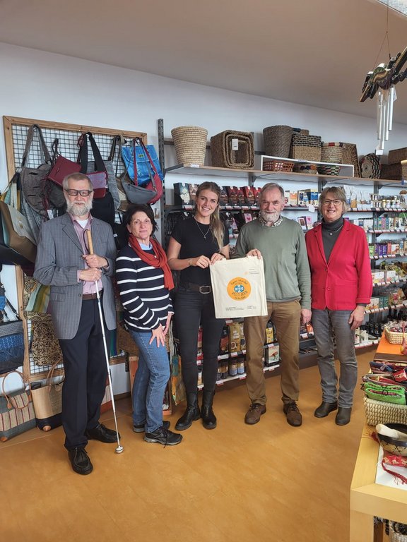 In einem kleinen Laden hängen im Hintergrund verschieden Verkaufsartikel. 3 Frauen und 2 Männer stehen nebeneinander und halte in der Mitte eine Baumwolltasche ins Bild. Die Personen lächeln in die Kamera, sind legere angezogen. Die Situation vermittelt eine Geschenkübergabe.