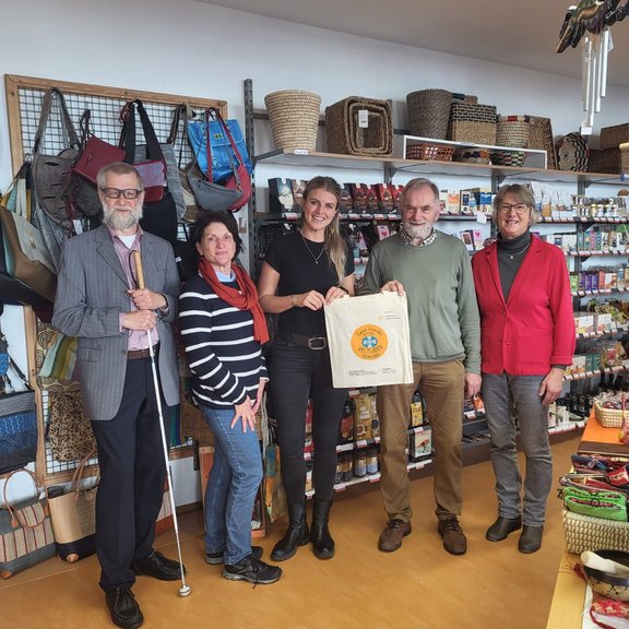 In einem kleinen Laden hängen im Hintergrund verschieden Verkaufsartikel. 3 Frauen und 2 Männer stehen nebeneinander und halte in der Mitte eine Baumwolltasche ins Bild. Die Personen lächeln in die Kamera, sind legere angezogen. Die Situation vermittelt eine Geschenkübergabe.