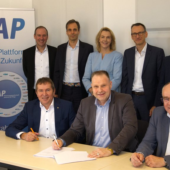 Männer und Frauen in Business-Kleidung sitzen und stehen an einem Tisch, halte teilweise einen Stift in der Hand. Hinten ist ein Roll-Up mit der Aufschrift "TAP" zu sehen. Die Szene vermittelt eine Vertragsunterzeichnung oder einen Abschluss.