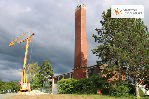 Industriegebäude, links ein gelber Kran in der Mitte ein gemauerter Kamin und rechts ein grüner Baum