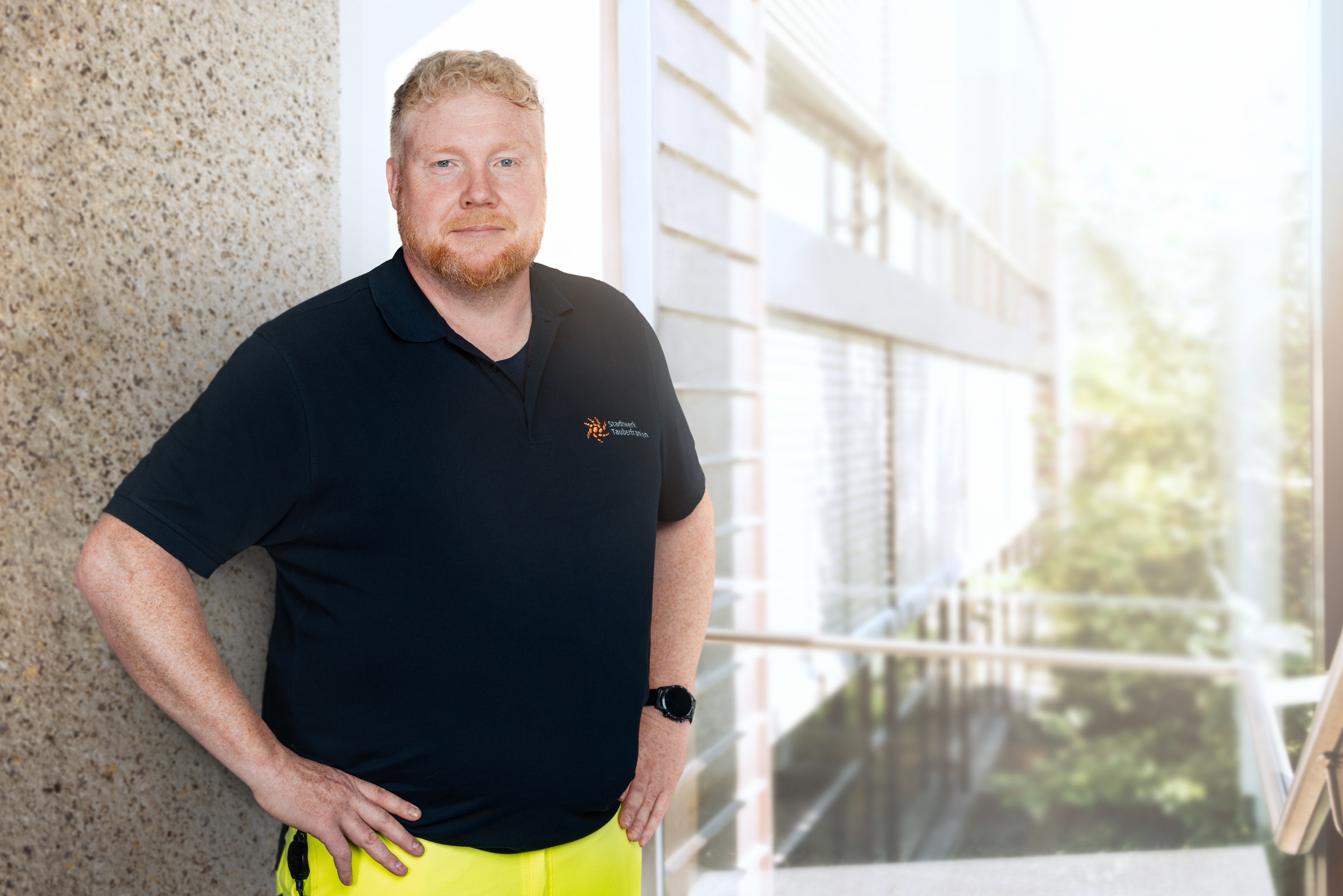 Strommonteur Ein Mann mit hellem Bart und kurzen, lockigen Haaren steht in einem modernen Gebäude an einer hellen Glasfassade. Er trägt ein dunkelblaues Polo-Shirt mit dem Logo des Stadtwerks Tauberfranken und leuchtend gelbe Arbeitshosen. Seine Haltung ist selbstbewusst, eine Hand ruht in der Hüfte. Die Szenerie wirkt freundlich und professionell.